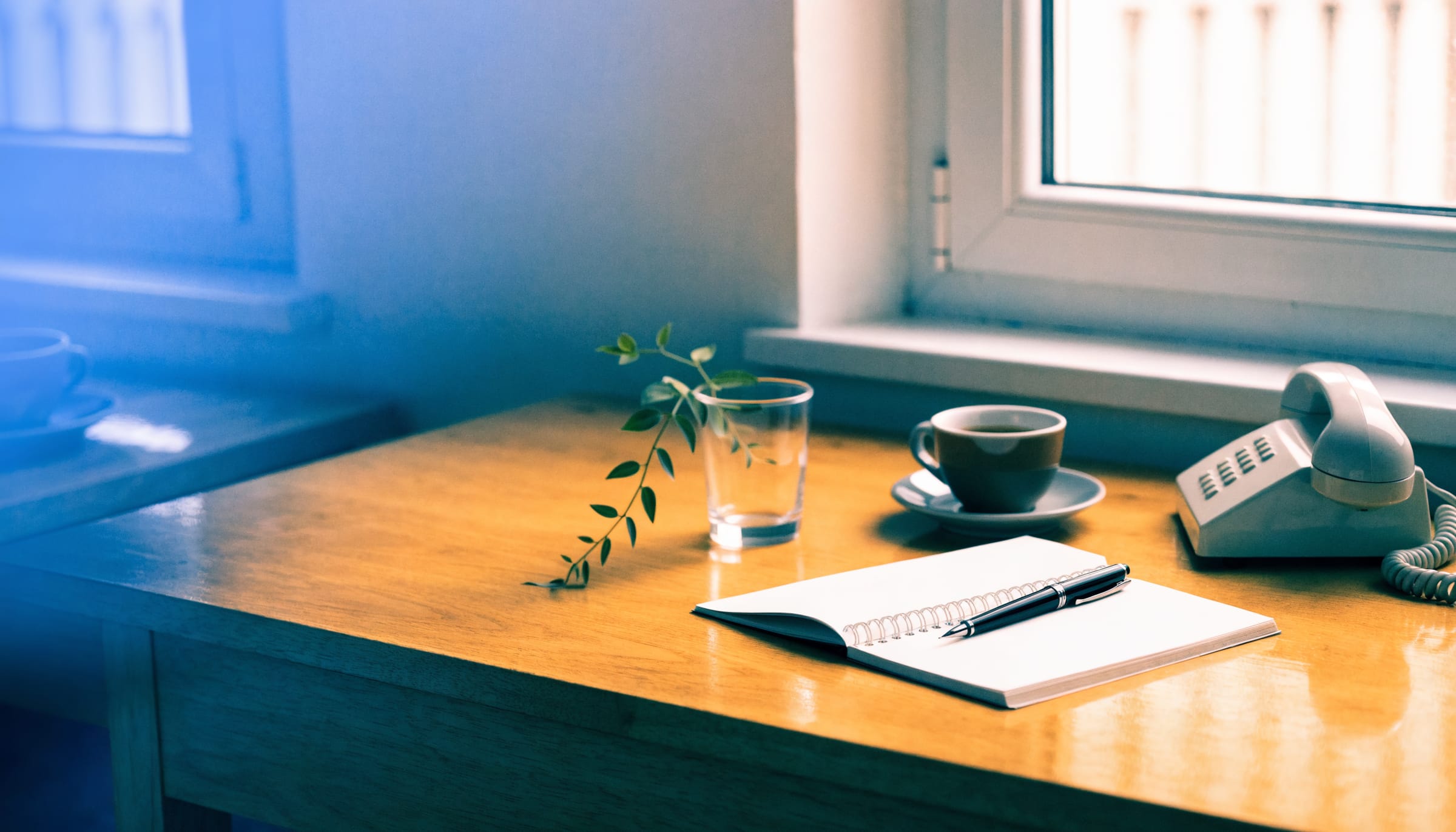A desk by a window with an open notebook, a pen, a cup of coffee and a cream telephone