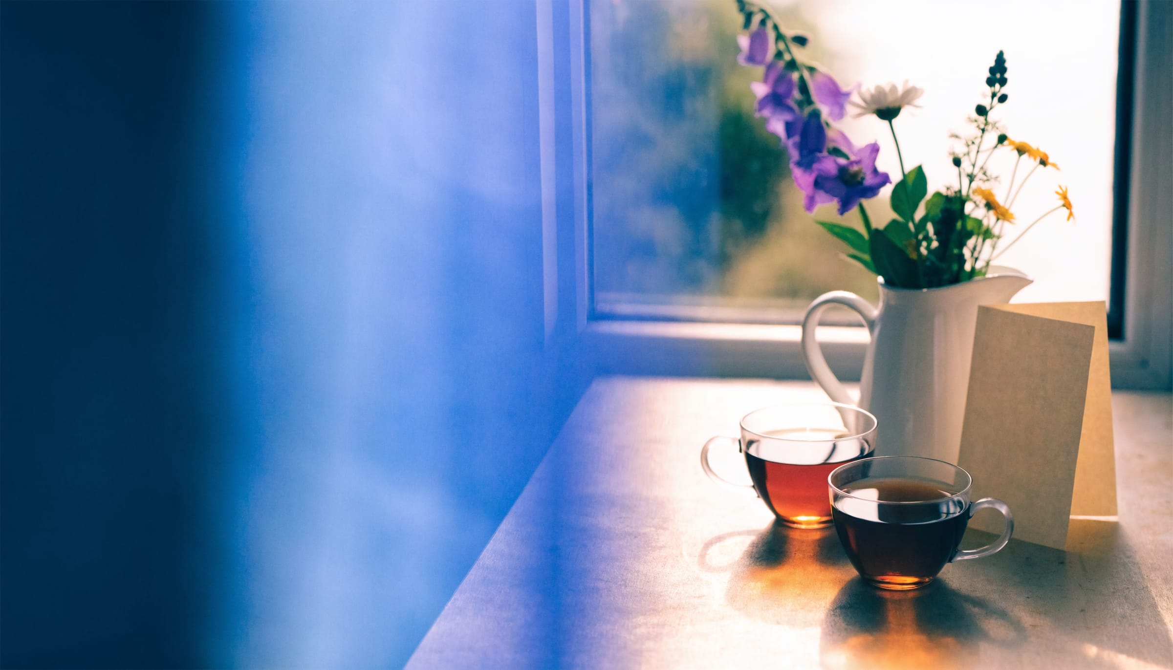 Two cups of tea, garden flowers in a jug and a blank card on a sunlit windowsill